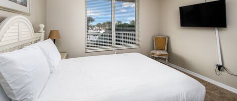 A hotel room with a large bed, a TV mounted on the wall, a window with blinds, a chair, and a lamp.