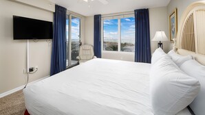 A bedroom with a large bed, a flat-screen TV mounted on the wall, a chair by the window, and a view of the beach. - IP 202: Gulf Dwelling -  Welcome to Your Beachfront Paradise! (Fort Walton Beach)