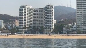 A high-rise hotel with multiple floors, a beach in the foreground, and a hilly landscape in the background.