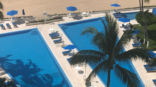 A large rectangular swimming pool with multiple lounge chairs and umbrellas, surrounded by palm trees and a sandy beach.