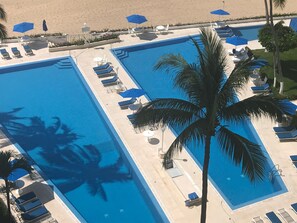 A large rectangular swimming pool with multiple lounge chairs and umbrellas, surrounded by palm trees and a sandy beach. - Beachfront Condo-Acapulco-Magnificent Views (Acapulco)