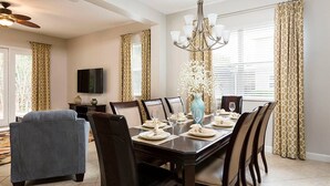 Uma sala de jantar com uma mesa de madeira escura posta para quatro pessoas, cercada por seis cadeiras. Um lustre pendurado sobre a mesa, e uma TV de tela plana fixada na parede.