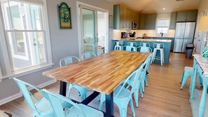 A dining area with a wooden table and light blue chairs, a kitchen with green cabinets, and a view of the outside through a sliding door. - Kaptain's Treasure | 5BR Carova Beach 4x4 OBX | Pvt Pool, Hot Tub & Game Rm (Carova)