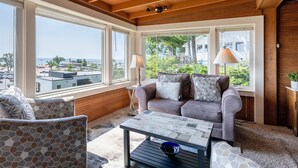 Ein gemütliches Wohnzimmer mit Sofa, Sessel und Couchtisch, großen Fenstern mit Blick auf das Meer und Gebäude sowie Holzbalkendecken.