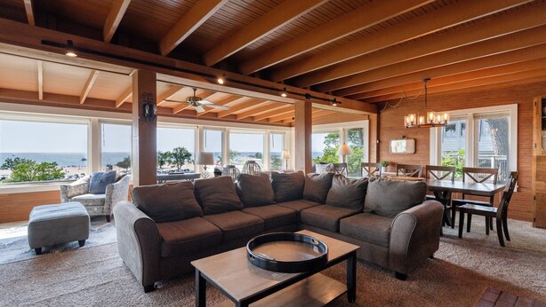 Ein geräumiges Wohnzimmer mit einer großen Eckcouch, einem Couchtisch und einem Essbereich mit Tisch und Stühlen. Große Fenster bieten Blick aufs Meer.