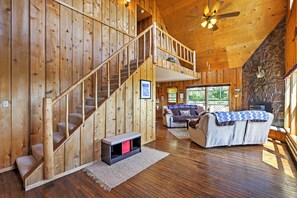 A wooden cabin interior with a staircase, a living area with a sofa and a fireplace, and a stone wall with a deer head carving.