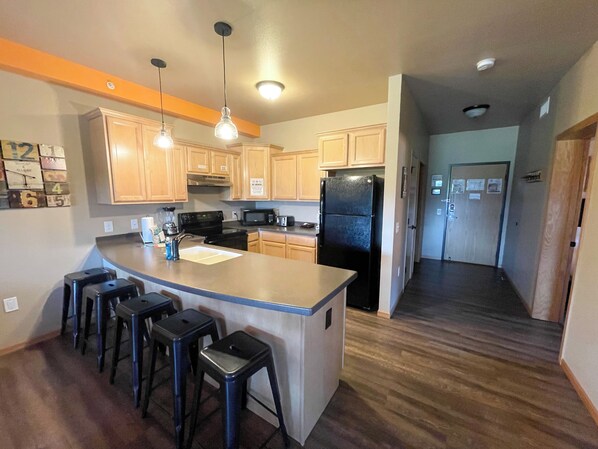 A kitchen with wooden cabinets, a black refrigerator, a microwave, and a sink. There are three stools at the island. - Two Bedroom Condo @ Spring Brook Resort (Wisconsin Dells)