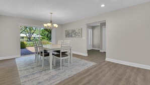 Uma sala de jantar ampla com mesa e cadeiras brancas, um lustre e vista para um jardim através de uma porta aberta.