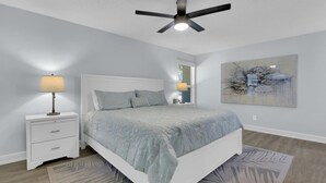 Quarto com cama, mesa de cabeceira, abajur, ventoinha no teto e uma obra de arte na parede.