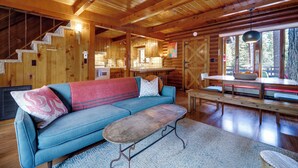Ein gemütliches Blockhaushaus-Interieur mit einer Holztreppe, einem blauen Sofa, einem Esstisch und Blick ins Freie.