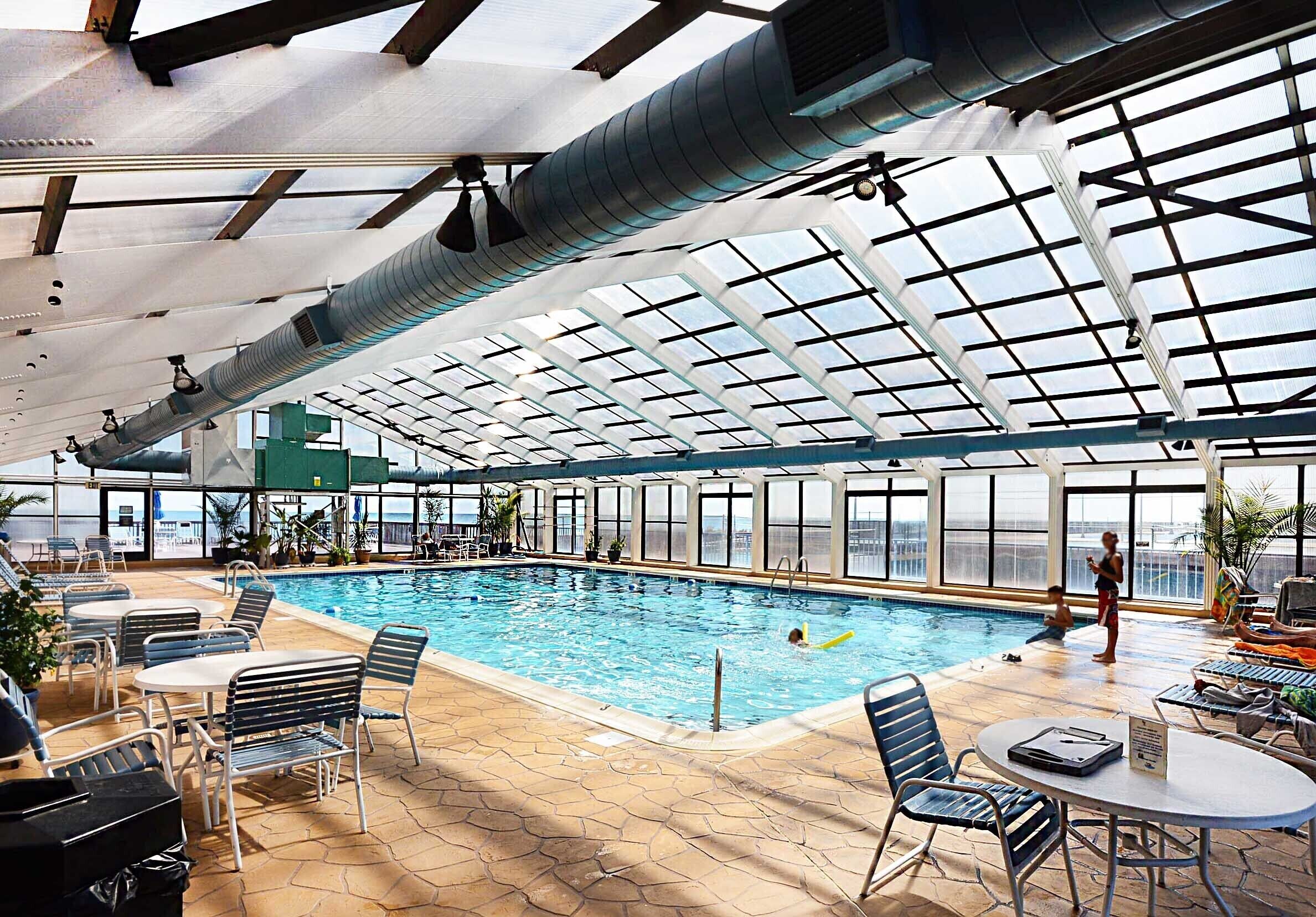 Ein Hallenbad mit klaren Glasdächern, Liegestühlen und Tischen.