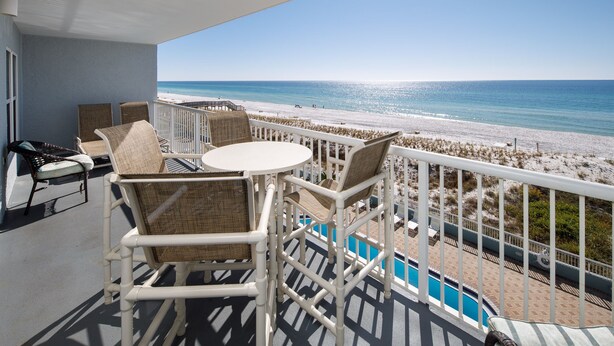 Un balcon avec des fauteuils en osier et une table, d’où l’on a vue sur une plage et une piscine.