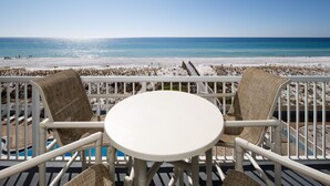 Un balcon avec une table blanche et deux chaises en osier, donnant sur une plage avec une vue dégagée sur l’océan.
