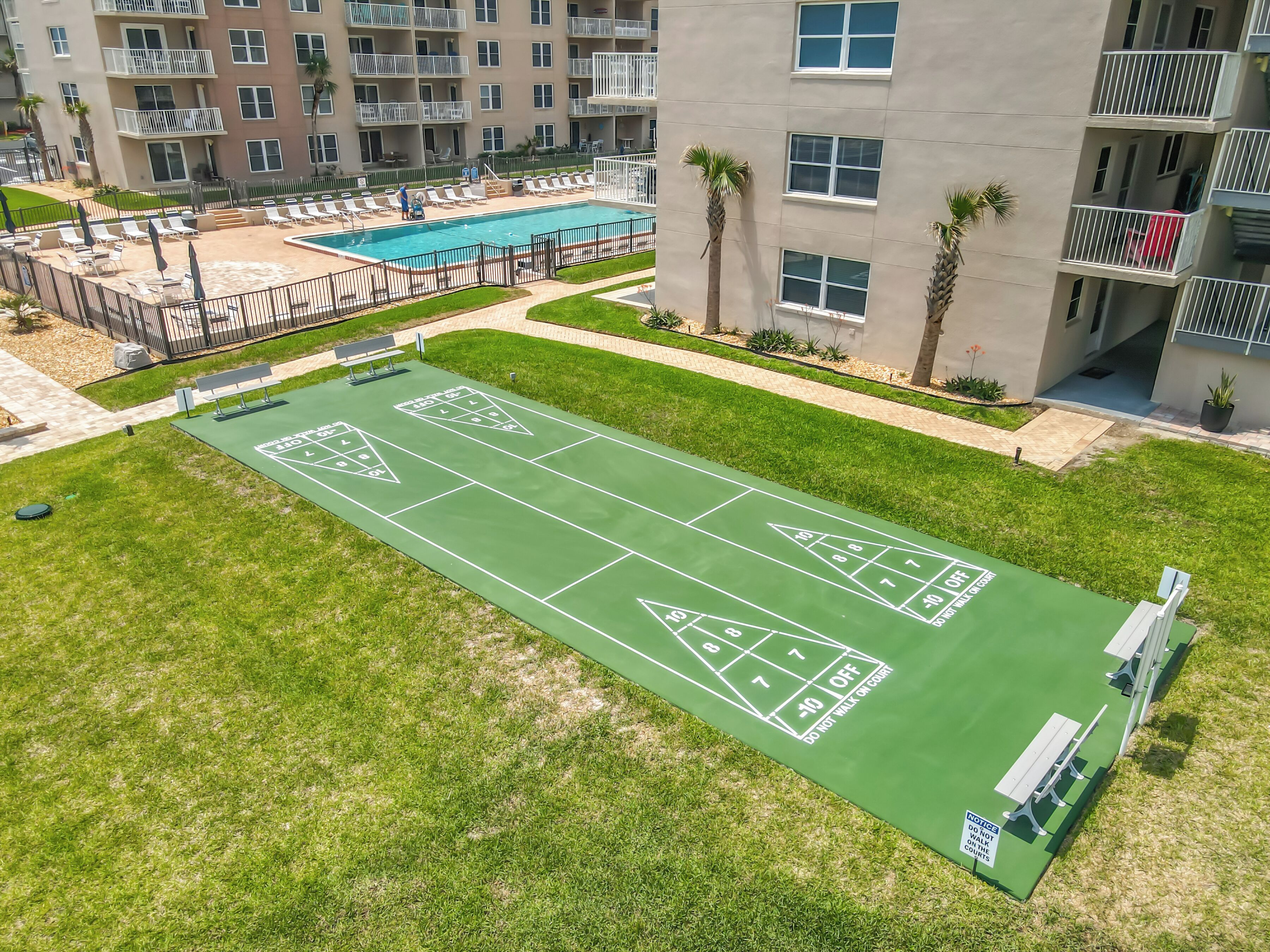 A green shuffleboard court surrounded by a pool area and a building with balconies.