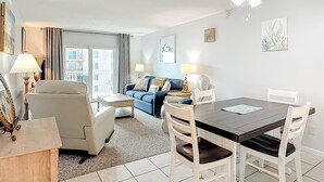 A living room with a dining table, white chairs, a blue sofa, and a ceiling fan. - Cute 2/2!Direct Oceanfront Complex on the No Drive Beach in NSB! (New Smyrna Beach)