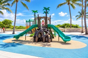 A playground with slides and climbing structures, surrounded by palm trees and a clear sky. - South of Fifth 1BR Condo - Heated Pool, Balcony & Walk to Beach (Miami Beach)