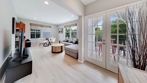 A modern living room with a sofa, coffee table, and dining area. There is a large window with blinds, a door leading to an outdoor patio, and a TV on a stand.