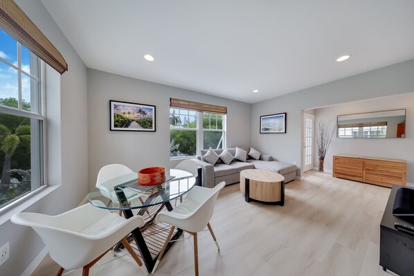 A modern living room with a glass dining table, white chairs, a grey sofa, and a wooden cabinet. - South of Fifth 1BR Condo - Heated Pool, Balcony & Walk to Beach (Miami Beach)
