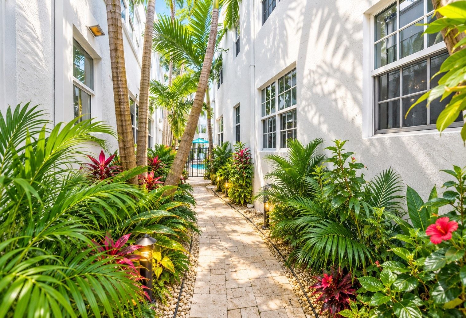 A narrow pathway flanked by white buildings with windows, surrounded by lush tropical plants and a clear blue sky.