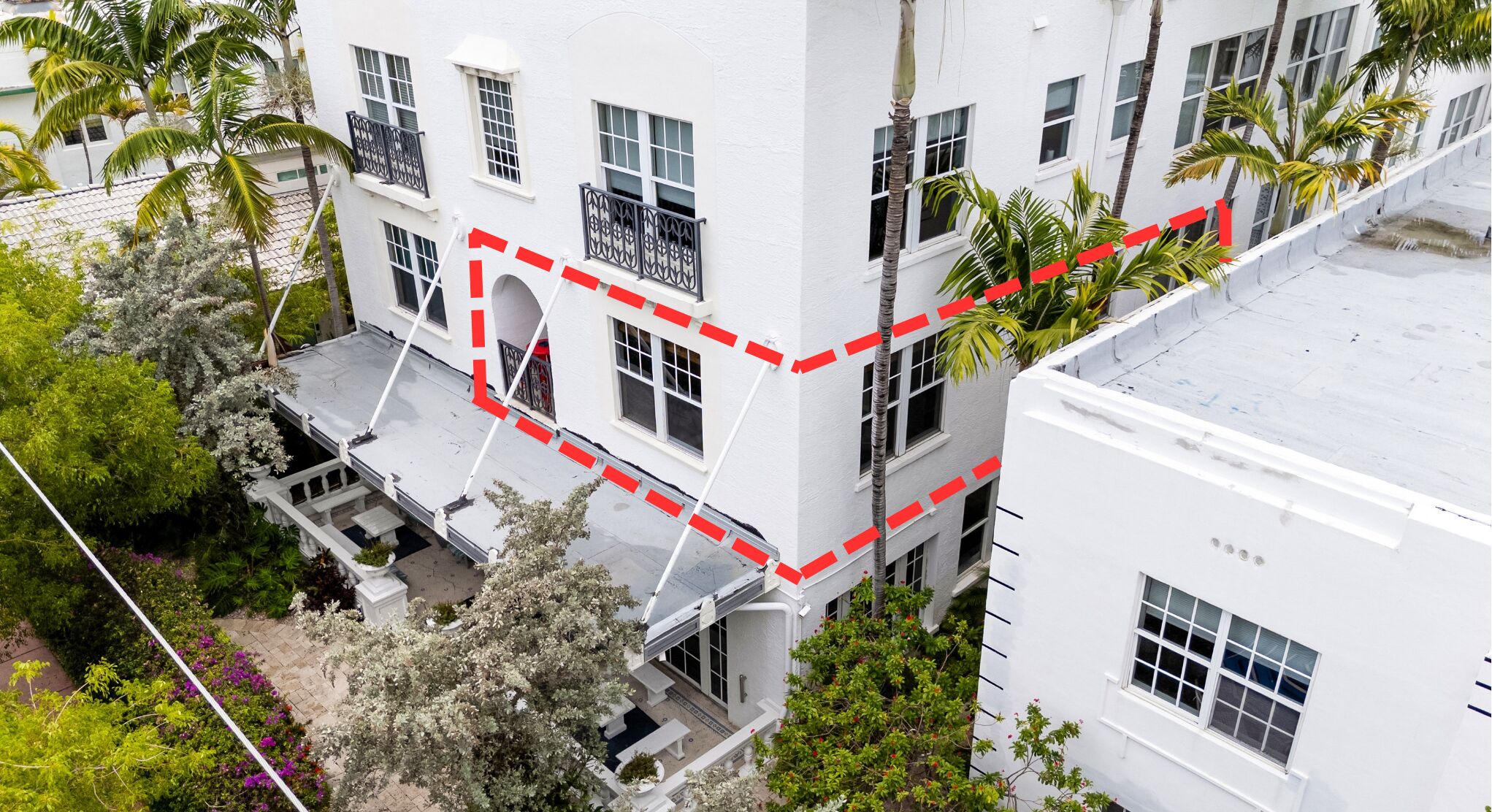 Un immeuble à plusieurs étages doté de balcons, de murs extérieurs blancs et entouré de verdure, notamment de palmiers et de plantes à fleurs.