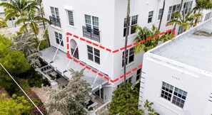 A multi-story building with balconies, white exterior walls, and surrounding greenery including palm trees and flowering plants. - South of Fifth 1BR Condo - Heated Pool, Balcony & Walk to Beach (Miami Beach)