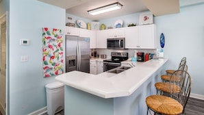 Una cocina moderna con armarios blancos, un refrigerador de acero inoxidable, un microondas y una estufa. Hay tres sillas de mimbre y un cubo de basura blanco.