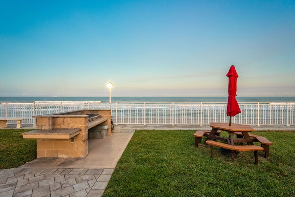 A picnic area with a table and benches, a red umbrella, and a view of the ocean. - Direct Oceanfront Condo with Extra-Large Balcony & Panoramic Views! 🌊 (Satellite Beach)