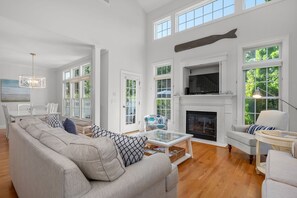 A spacious living room with a fireplace, large windows, and a high ceiling. - Captain's Row: Pool, Tennis & Chatham Charm (Chatham)