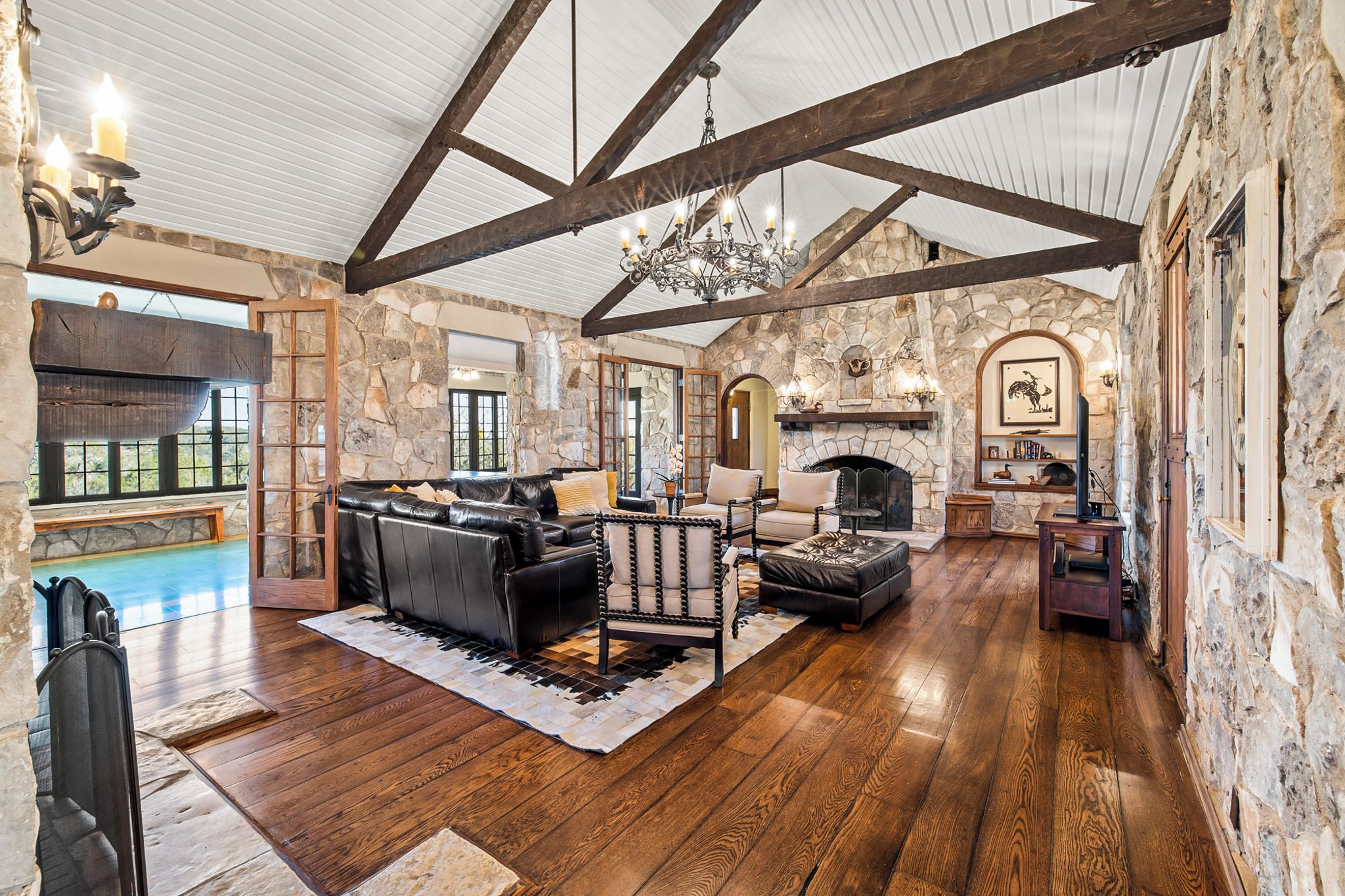 A spacious living room with a stone fireplace, wooden beams, and a large window overlooking a pool.