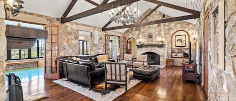 A spacious living room with a stone fireplace, wooden beams, and a large window overlooking a pool.