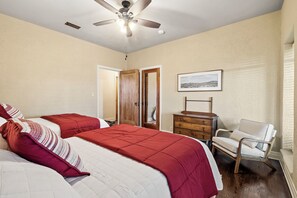 Une chambre avec deux lits, un ventilateur de plafond, une commode, une chaise et un tableau encadré au mur.