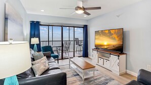 A living room with a sofa, coffee table, TV, and a balcony with a view of the sunset.