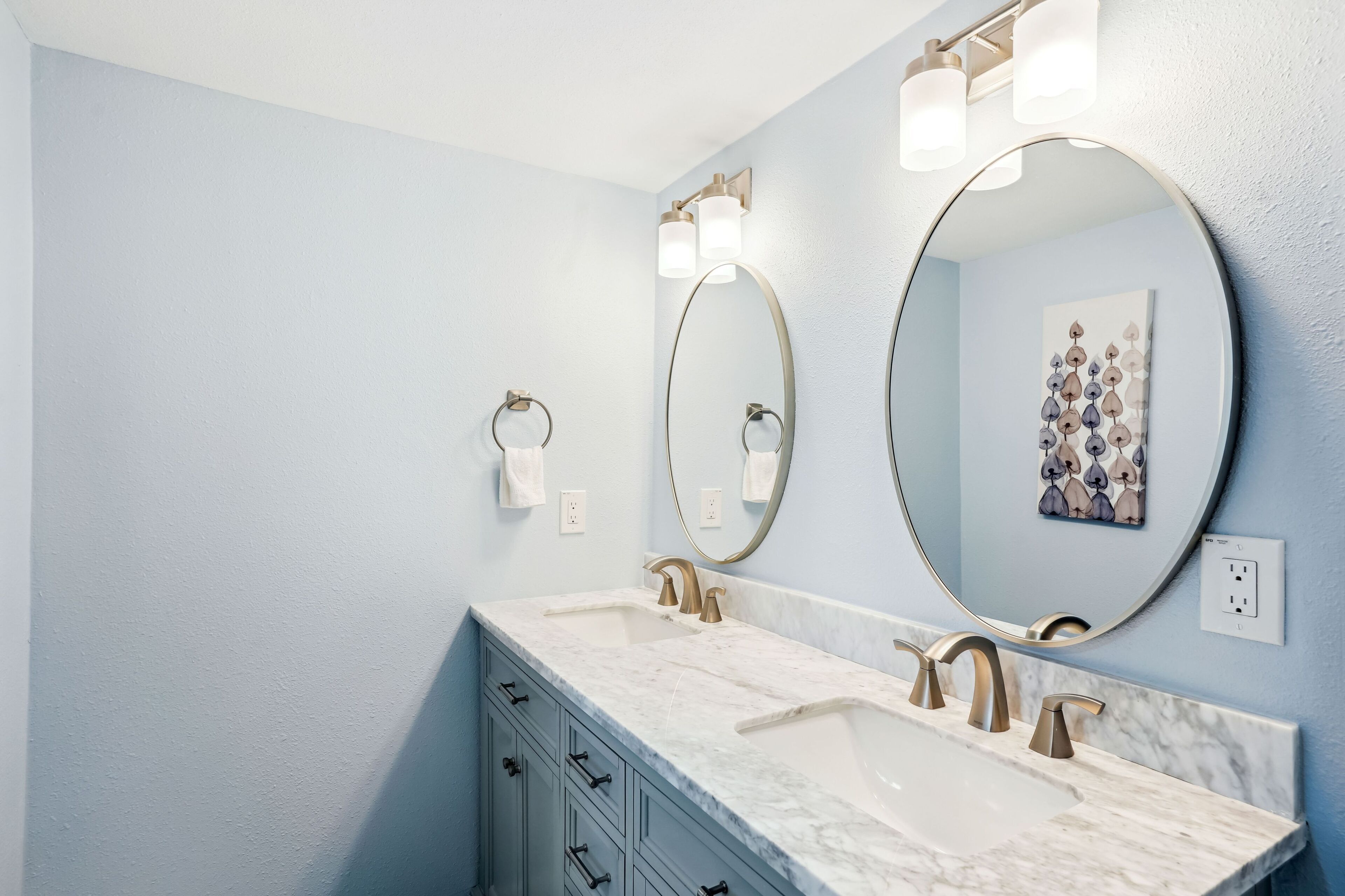 A bathroom with a double sink, gold faucets, a large round mirror, and a towel ring on the wall.