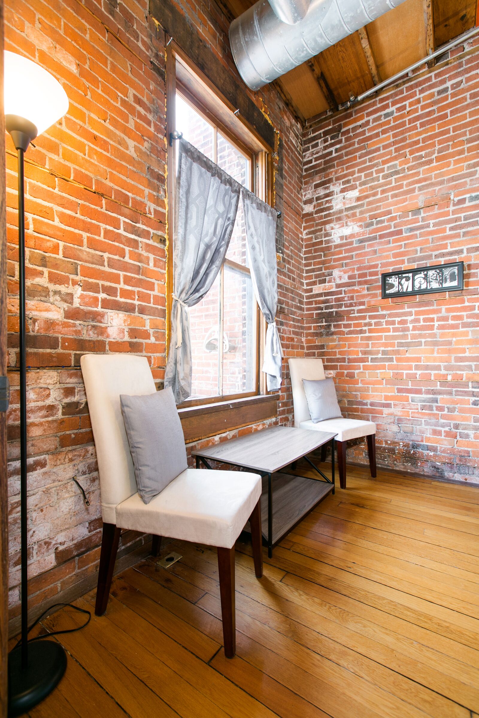 A room with two white cushioned chairs, a wooden floor, a brick wall, and a window with curtains.