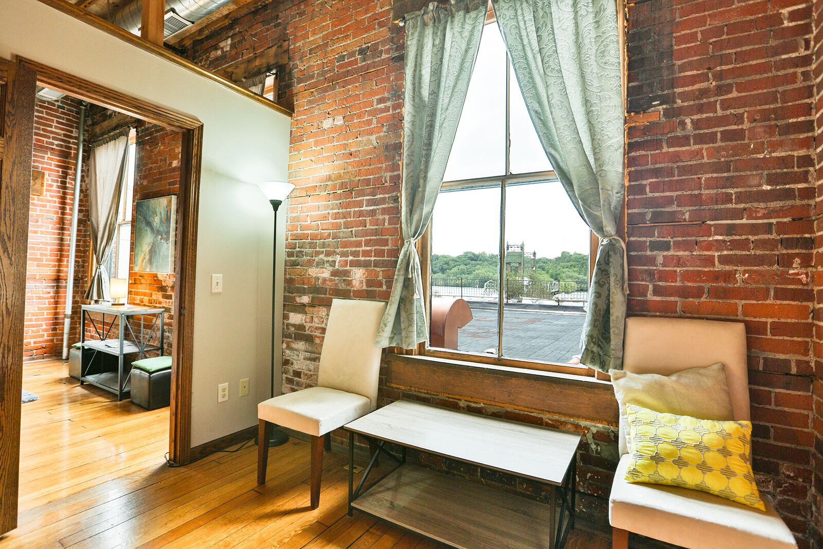 A room with a brick wall, a large window with curtains, a bench, and a table with a lamp.