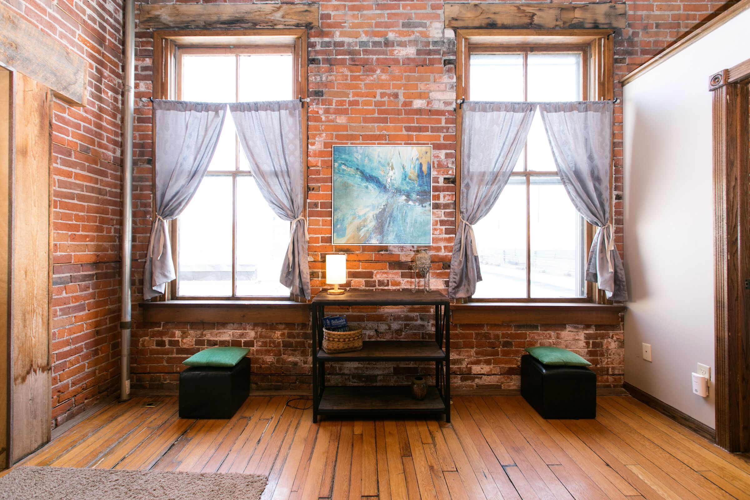 Una habitación con pared de ladrillo, dos ventanas con cortinas, suelo de madera, un mueble de televisión negro con un cuadro, y dos otomanas verdes.