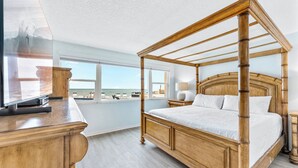 A four-poster bed with a wooden frame, a television on a wooden stand, and a view of the beach through large windows. - Perfect for Families - Oceanfront Ground Floor Condo w/ Large Heated Pool! 🏖️ (Satellite Beach)