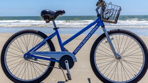 Un vélo bleu avec un panier à l’avant, garé sur une plage de sable, avec l ’océan en arrière-plan.