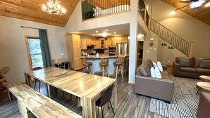 A spacious living area with a wooden dining table, bar stools, a sofa, and a staircase leading to an upper level.