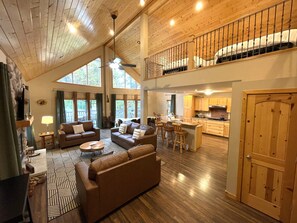 Un espacio habitable amplio con techo alto, pisos de madera y una cocina al fondo.