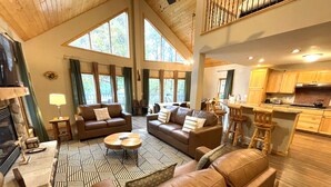 A spacious living room with leather sofas, a coffee table, and a patterned rug. There is a fireplace, a dining area, and a kitchen with wooden cabinets.