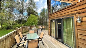 A wooden deck with outdoor furniture, a glass door entrance, and a view of trees and sky.