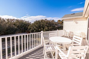 A white outdoor patio with a round table and chairs, a white fence, and a view of trees and a building in the background.