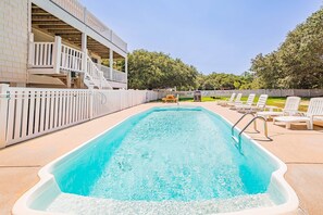 A rectangular swimming pool with a white deck surrounding it, a wooden deck area with a table and chairs, and a white fence enclosing the area.