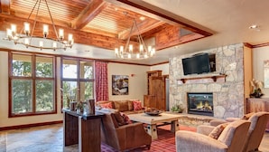 A spacious living room with a stone fireplace, wooden ceiling beams, and a large TV mounted on the wall. - Ski-In/Ski-Out! Hot tub access, sleeps 9, Heated garage parking! (Breckenridge)