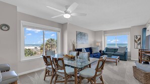 A spacious living room with a glass dining table surrounded by wicker chairs, a blue sofa, and a ceiling fan. - Completely Renovated Oceanfront Unit in Quaint Surfside Beach! SC205b (Surfside Beach)