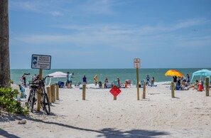A beach with a sign indicating beach access is closed, a no parking sign, and people relaxing under umbrellas.