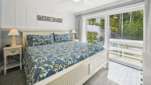 Une chambre avec un lit, des tables de chevet, un ventilateur de plafond, une fenêtre donnant sur un paysage verdoyant et une pancarte indiquant « CABANE DE PLAGE ».