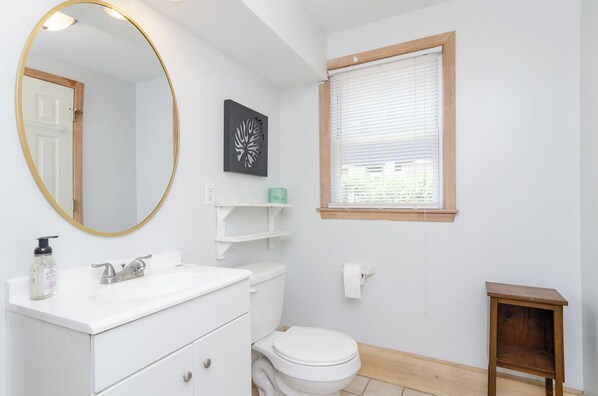 Un baño con un lavamanos blanco, un inodoro, un espejo con marco dorado, una ventana con persianas y un taburete de madera.