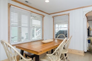Un comedor con mesa de madera y sillas blancas, ventanas grandes con persianas y vista a la calle.